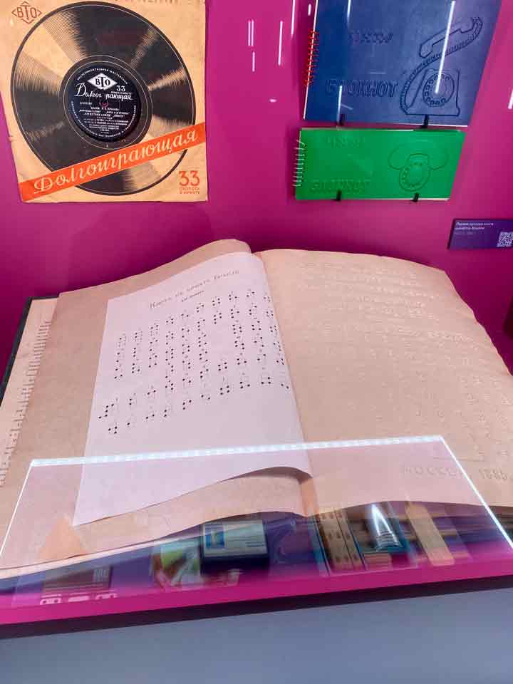 Тифлокомментарий к фото 1: Витрина с раскрытой книгой, напечатанной шрифтом Брайля, виниловым диском и двумя блокнотами с рельефными изображениями старых стационарных телефонов.