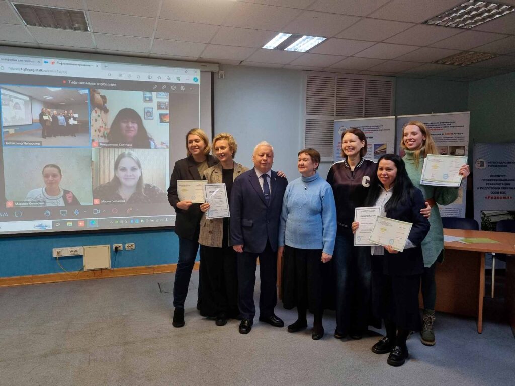 Тифлокомментарий 1: Общая фотография выпускников программы: На экране – дистанционные участники: Мария Белькова, Светлана Мещерякова, Марина Копысова. Справа от экрана – тифлокомментаторы получившие удостоверения очно: Ирина Безрукова, Анна Цанг, Валерия Сатлер, Гелюся Закирова, Янина Исаичкина вместе с авторами российской концепции тифлокомментирования Сергеем Николаевичем и Ольгой Павловной Ваньшиными.