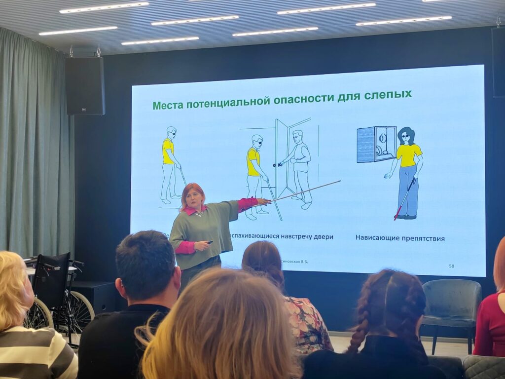 Лекторий. Перед экраном с презентацией преподаватель - В.В. Коновалова. Левой рукой она держит указку, направленную на экран. На нём надпись "места потенциальной опасности для слепых" и несколько демонстрационных изображений.