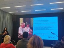 Тифлокомментарий: Лекторий. В центре, на экране, слайд презентации. На слайде фотография разработчиков отечественной концепции тифлокомментирования С.Н. и О.П. Ваньшиных и справочная информация. Слева за кафедрой М.А. Щербакова. Она говорит в микрофон. Перед ней несколько рядов слушателей.