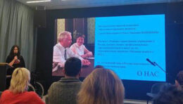 Тифлокомментарий: Лекторий. В центре, на экране, слайд презентации. На слайде фотография разработчиков отечественной концепции тифлокомментирования С.Н. и О.П. Ваньшиных и справочная информация. Слева за кафедрой М.А. Щербакова. Она говорит в микрофон. Перед ней несколько рядов слушателей.