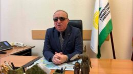 Тифлокомментарий к фотографии. На фотографии в своём кабинете за рабочим столом президент ВОС Владимир Васильевич Сипкин. Он в деловом костюме, руки на столе, сложены замком. За ним флаг Всероссийского общества слепых