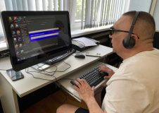 Тифлокомментарий к 1 фотографии: участник специальной военной операции за компьютерным рабочим местом: на нём наушники, он печатает на клавиатуре обеими руками.