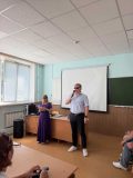 Тифлокомментарий: На фотографии — заместитель генерального директора по информационным технологиям Института «Реакомп» Е.Е. Арнапольский с микрофоном в правой руке. У него светлые волосы и тёмные очки. Он одет в светлую рубашку, чёрные брюки и белые кроссовки. Слева от него стоит организатор мероприятия. На ней длинное фиолетовое платье, светлые волосы спадают на плечи. Её взгляд устремлен на экран планшета, который она держит в руках. Перед спикерами за столами - участники мероприятия. На заднем плане — большой белый проекторный экран.