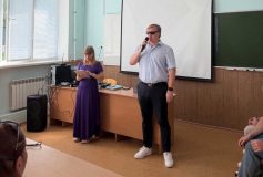 Тифлокомментарий: На фотографии — заместитель генерального директора по информационным технологиям Института «Реакомп» Е.Е. Арнапольский с микрофоном в правой руке. У него светлые волосы и тёмные очки. Он одет в светлую рубашку, чёрные брюки и белые кроссовки. Слева от него стоит организатор мероприятия. На ней длинное фиолетовое платье, светлые волосы спадают на плечи. Её взгляд устремлен на экран планшета, который она держит в руках. Перед спикерами за столами - участники мероприятия. На заднем плане — большой белый проекторный экран.